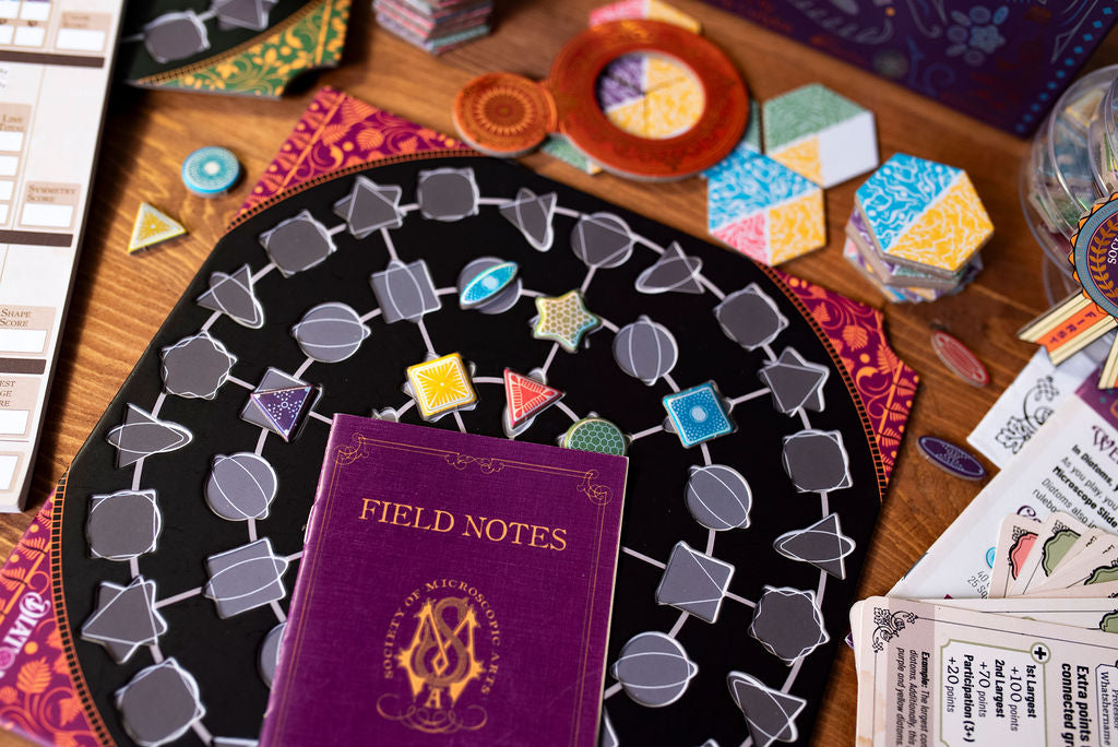A close-up of various Diatoms game components including a purple Field Notes book on top of a player board, water tiles, and other pieces surround the photo perimeter.