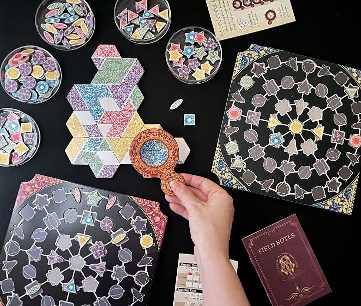 A top-down photo of the components of Diatoms the board game. 5 petri dishes of colorful algae tiles are on the upper left. The center shows a display of connected water tiles with the lens held over a tile intersection. Two player boards are visible on the bottom and right side of the image and in the bottom right corner is a player reference Field Notes book