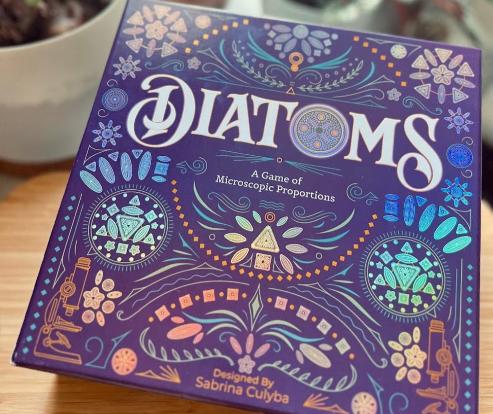 A photo of the Diatoms board game box, tilted on a table showing off the holographic shimmer of the cover. In the background are blurred hints of potted plants