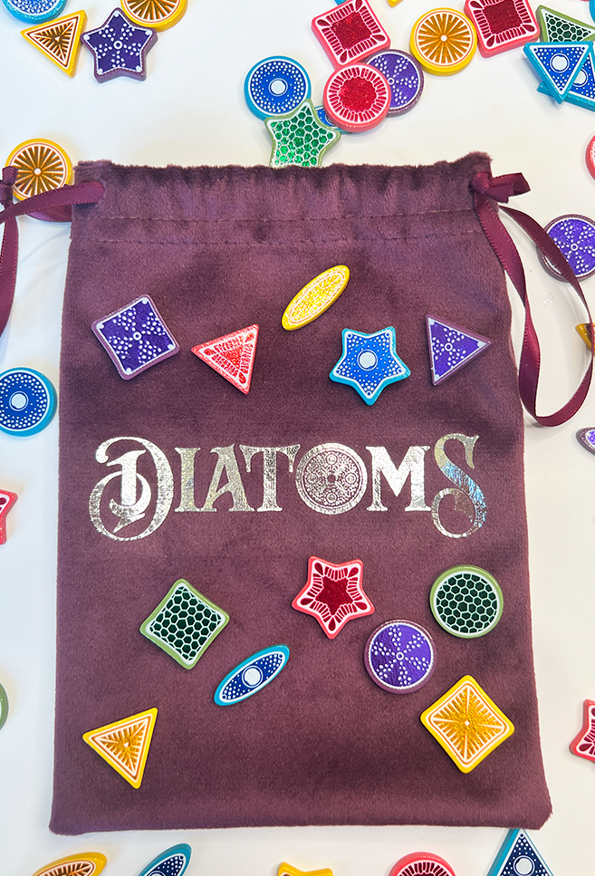 Photo of the deluxe wooden tiles add-on for Diatoms the board game. In the center is the purple velvet draw bag with silver DIATOMS logo. Strewn around and on top of the bag are blue, pink, yellow, purple, and green wooden diatom tiles.