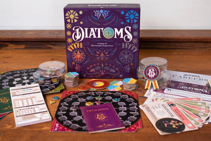 A photo of Diatoms the board game with the box standing up on a wooden table and all components arrayed around it.